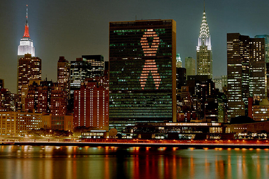 The UN Secretariat Building is lit as a Red Ribbon with the East River in the foreground.